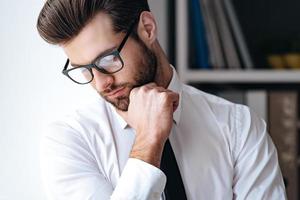 moderner Geschäftsmann. Porträt eines hübschen jungen Geschäftsmannes mit Brille, der nach unten schaut und die Hand am Kinn hält, während er im Büro steht foto