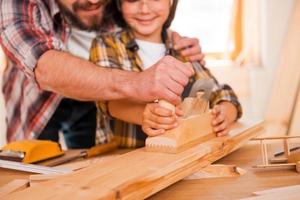 Fähigkeiten mit seinem Sohn teilen. Nahaufnahme eines lächelnden jungen männlichen Tischlers, der seinem Sohn beibringt, in seiner Werkstatt mit Holz zu arbeiten foto