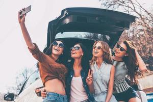 Deshalb machen wir Roadtrips mit vier schönen jungen, fröhlichen Frauen, die mit einem Lächeln ein Selfie machen, während sie in der Nähe des Kofferraums stehen foto