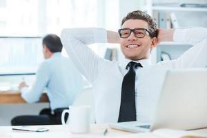 zufrieden mit der geleisteten Arbeit. Fröhlicher Geschäftsmann in Abendkleidung, der den Kopf in den Händen hält, während er an seinem Schreibtisch im Büro sitzt foto