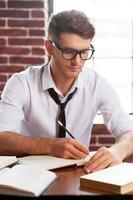 auf die Arbeit konzentriert. Selbstbewusster junger Mann in Hemd und Krawatte, der etwas in den Notizblock schreibt, während er an seinem Arbeitsplatz sitzt foto