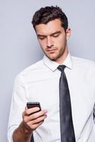 Geschäftsnachricht eingeben. Selbstbewusster junger Mann in Hemd und Krawatte, der ein Handy hält und es betrachtet, während er vor grauem Hintergrund steht foto