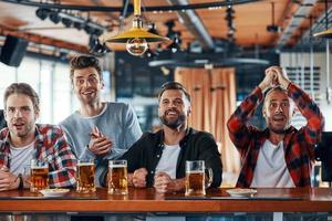 Jubelnde junge Männer in Freizeitkleidung, die Bier trinken und Sportspiele beobachten, während sie in der Kneipe sitzen foto