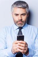 Geschäftsnachricht eingeben. Selbstbewusster reifer Mann in Hemd und Krawatte, der ein Handy hält und es betrachtet, während er vor grauem Hintergrund steht foto