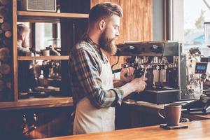 den besten Kaffee in dieser Stadt zu machen. Seitenansicht eines jungen bärtigen Mannes in Schürze, der Kaffee kocht, während er im Café steht foto