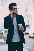 Ich werde rechtzeitig sein. hübscher junger Mann in eleganter Freizeitkleidung, der eine Kaffeetasse trägt und mit dem Handy spricht, während er die Straße entlang geht foto