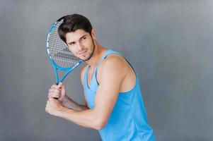 Tennisspieler. selbstbewusster junger muskulöser Mann, der Tennisschläger hält, während er vor grauem Hintergrund steht foto