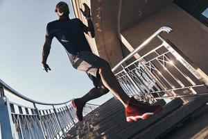 sich anstrengen. Volle Länge des jungen afrikanischen Mannes in Sportkleidung, der die Treppe hinunterläuft, während er im Freien trainiert foto
