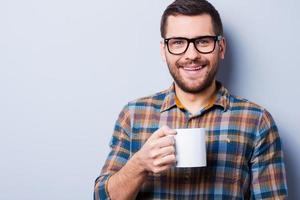 Zeit, eine Bremse zu haben. hübscher junger Mann trinkt Kaffee und schaut in die Kamera, während er vor grauem Hintergrund steht foto