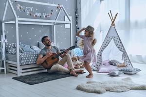 zusammen Spaß haben. Junger Vater spielt Gitarre für seine kleine Tochter und lächelt, während er Freizeit zu Hause verbringt foto