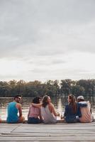 tolle Zeit mit Freunden. Rückansicht junger Menschen in Freizeitkleidung, die reden und lächeln, während sie auf dem Pier sitzen foto