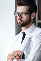 Analyse von Mediennachrichten. nachdenklicher junger geschäftsmann mit brille, der aufgerollte zeitung hält und wegschaut, während er im büro steht foto