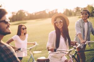 gute zeit mit besten freunden verbringen. glückliche junge leute, die ihre fahrräder schieben, während sie zusammen durch grünes gras gehen foto