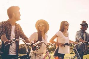 Qualitätszeit mit Freunden verbringen. fröhliche junge leute, die ihre fahrräder schieben, während sie gemeinsam durch grünes gras im freien gehen foto