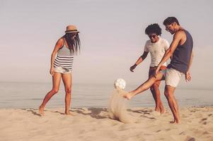 Freunde spielen am Strand. Drei fröhliche junge Leute spielen mit Fußball am Strand mit Meer im Hintergrund foto