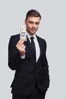 Geld verdienen. Gut aussehender junger Mann in vollem Anzug, der lächelt und eine Papierwährung hält, während er vor grauem Hintergrund steht foto
