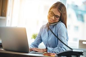 bis morgen fröhliche junge schöne frau mit brille, die am telefon spricht und einen laptop mit einem lächeln benutzt, während sie an ihrem arbeitsplatz sitzt foto