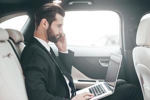 beschäftigt auf der Arbeit. Selbstbewusster junger Geschäftsmann, der an seinem Laptop arbeitet und telefoniert, während er im Auto sitzt foto