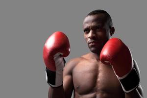 selbstbewusster Boxer. Selbstbewusster junger afrikanischer Boxer in Boxhandschuhen, der wegschaut, während er isoliert auf grau steht foto