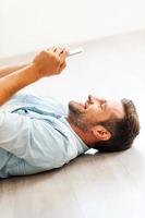 Zeit zum Entspannen nehmen. Seitenansicht eines jungen Mannes, der ein Handy hält und lächelt, während er in seiner Wohnung auf dem Boden liegt foto