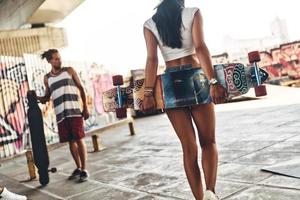 verliebt ins skaten. Rückansicht einer schönen jungen Frau, die ein Skateboard hinter ihrem Rücken hält, während sie mit Freunden im Skatepark im Freien rumhängt foto