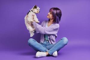 Fröhliche junge Frau, die kleinen Hund trägt und lächelt, während sie vor violettem Hintergrund sitzt foto