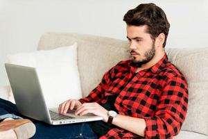 der bequemste Arbeitsplatz. ernsthafter junger Mann, der am Laptop arbeitet, während er auf dem Sofa sitzt foto