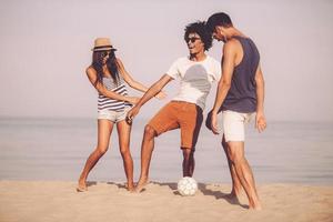 Spaß am Strand. Drei fröhliche junge Leute spielen mit Fußball am Strand mit Meer im Hintergrund foto