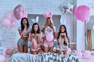 Jung und schön. Vier verspielte junge Frauen in Hasenohren, die ein Gesicht machen und gestikulieren, während sie auf dem Bett sitzen foto