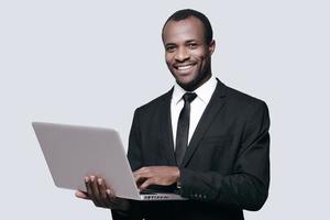 Erfolg erzielt hübscher junger afrikanischer Mann in Abendkleidung, der am Laptop arbeitet, während er vor grauem Hintergrund steht foto