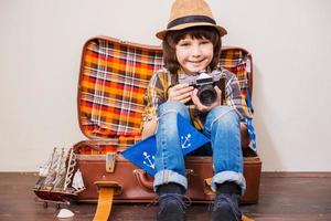 Sagen Sie Käse kleiner Junge in Kopfbedeckungen, der Kamera hält und lächelt, während er im Koffer vor braunem Hintergrund sitzt foto