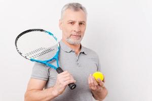 fertig zu spielen. Selbstbewusster älterer Mann mit grauem Haar, der Tennisschläger und Ball hält, während er vor weißem Hintergrund steht foto