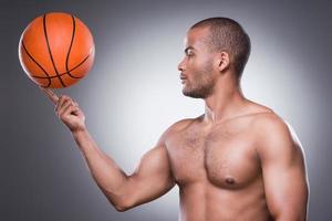 auf ein Spiel warten. Seitenansicht eines jungen, hemdlosen Afrikaners, der einen Basketballball am Finger trägt, während er vor grauem Hintergrund steht foto