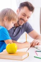 Sohn bei den Schulaufgaben helfen. Seitenansicht des fröhlichen jungen Vaters, der seinem Sohn bei den Hausaufgaben hilft und lächelt, während er zusammen mit grünem Apfel im Vordergrund am Tisch sitzt foto