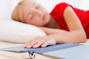 müde vom Lesen. süßes kleines Mädchen, das schläft, während es im Bett liegt und die Hand am Buch hält foto