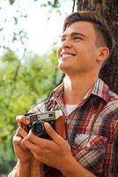 glücklicher Fotograf. hübscher junger Mann, der eine Vintage-Kamera hält und lächelt, während er sich an den Baum lehnt foto