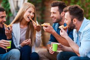 so leckere Gruppe fröhlicher junger Leute, die lächeln und Pizza essen, während sie im Freien sitzen foto