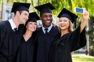 Einen glücklichen Moment festhalten. Vier Hochschulabsolventen in Abschlusskleidern, die nahe beieinander stehen und Selfie machen foto