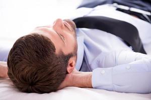 totale Entspannung. Rückansicht eines jungen Mannes in Hemd und Krawatte, der beim Schlafen im Bett die Hände hinter dem Kopf hält foto