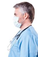 überzeugter Chirurg. Seitenansicht des reifen Arztes mit grauem Haar in OP-Maske, der in die Kamera blickt, während er isoliert auf Weiß steht foto