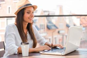 Nutzung des kostenlosen WLANs. Schöne junge Frau mit flippigem Hut, die am Laptop arbeitet und lächelt, während sie im Freien sitzt foto