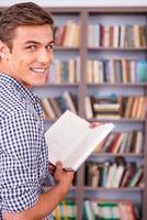 fröhlicher Bücherwurm. Rückansicht eines glücklichen jungen Mannes, der ein Buch hält und über die Schulter schaut, während er gegen ein Bücherregal steht