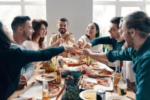 Beeilen Sie sich, um das beste Stück zu finden Gruppe junger Leute in Freizeitkleidung, die Pizza pflücken und lächeln, während sie drinnen eine Dinnerparty haben foto