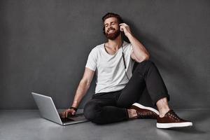 Musik genießen. Hübscher junger Mann, der Musik hört und Computer benutzt, während er auf dem Boden vor grauem Hintergrund sitzt foto
