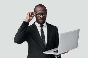 Ernster junger afrikanischer Mann in Abendkleidung, der mit dem Computer arbeitet, während er vor grauem Hintergrund steht foto