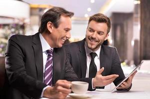 erfolgreiches Projekt. Zwei fröhliche Geschäftsleute in Abendgarderobe diskutieren etwas und lächeln, während einer von ihnen auf ein digitales Tablet zeigt foto