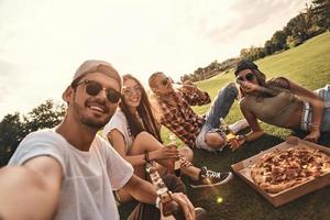 Selbstporträt von jungen Leuten in Freizeitkleidung, die lächeln, während sie Pizza und Bier im Freien genießen foto