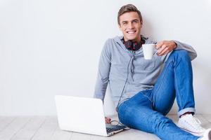 sich glücklich und entspannt fühlen. Schöner junger Mann mit Kopfhörern, der eine Kaffeetasse hält und lächelt, während er mit einem Laptop in seiner Nähe auf dem Boden sitzt foto