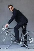 umweltschonender Transport. in voller Länge vom überzeugten jungen Geschäftsmann, der auf Fahrrad sitzt und Kamera beim Radfahren gegen grauen Hintergrund betrachtet foto