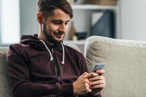 freut sich über sein neues Smartphone. Fröhlicher junger Mann, der sein Smartphone mit einem Lächeln benutzt, während er zu Hause auf der Couch sitzt foto